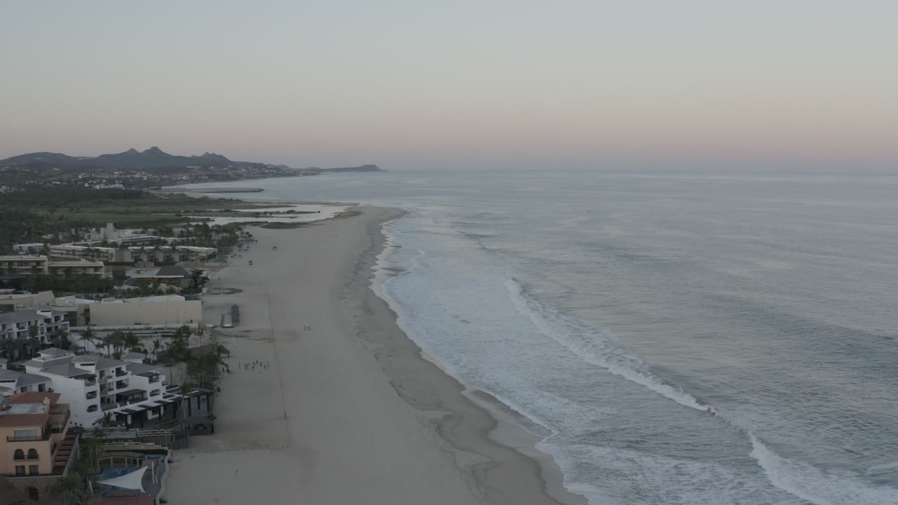 Opening aerial frame over a Los Cabos beach, with beachfront buildings at lower left, pale sand curving along gray-blue ocean, distant hills, and no clearly visible people.