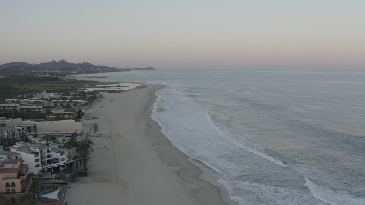 Aerial view in Los Cabos looking down the shoreline, with low waves rolling toward sand, white coastal buildings at lower left, distant headlands, and no clearly visible people.