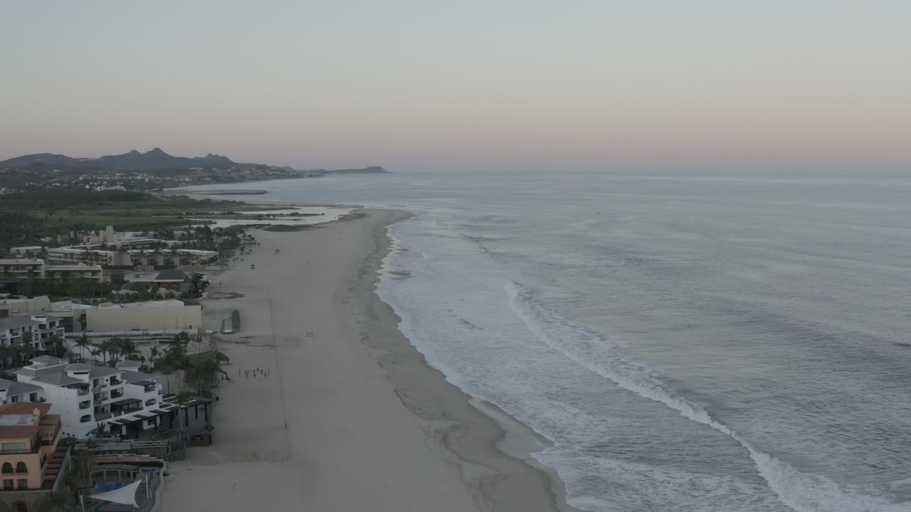 Wide coastal aerial with ocean to the right and beachfront structures to the left.
