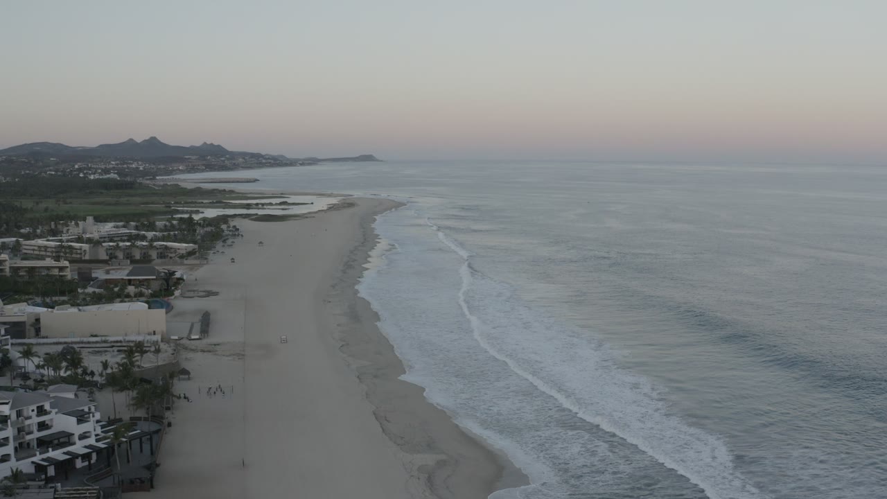 Los Cabos shoreline from above, with diagonal surf lines, broad empty sand, resort-area buildings along the left edge, low hills in the background, and no visible people.
