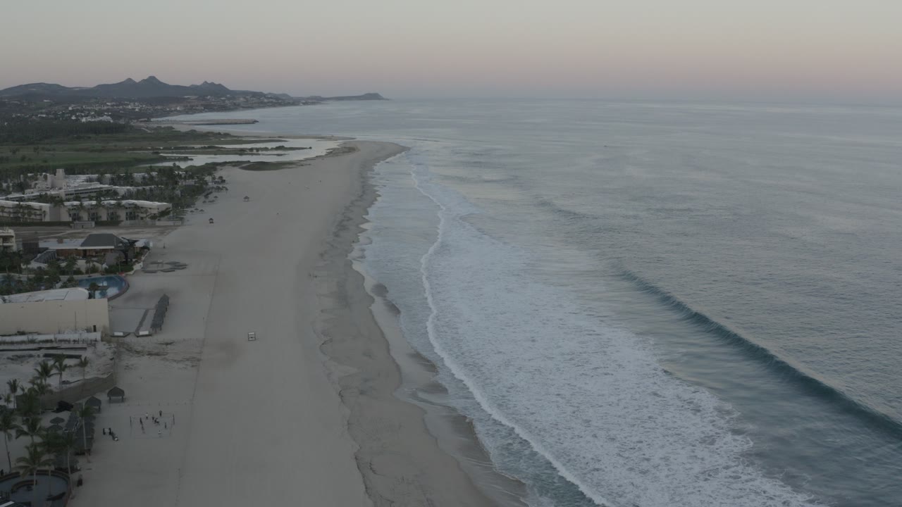 High aerial view over a Los Cabos beach with gray-blue ocean on the right, white surf lines, resort buildings at lower left, distant hills, and tiny people visible on the sand.