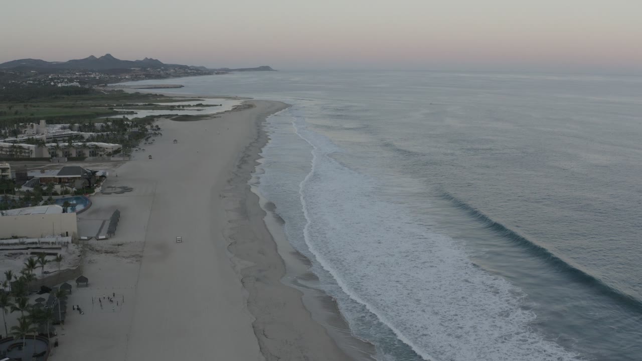 Aerial Los Cabos shoreline view with a long diagonal beach, ocean swells breaking near shore, resort structures at the lower left edge, distant hills, and tiny people visible near the beach access.