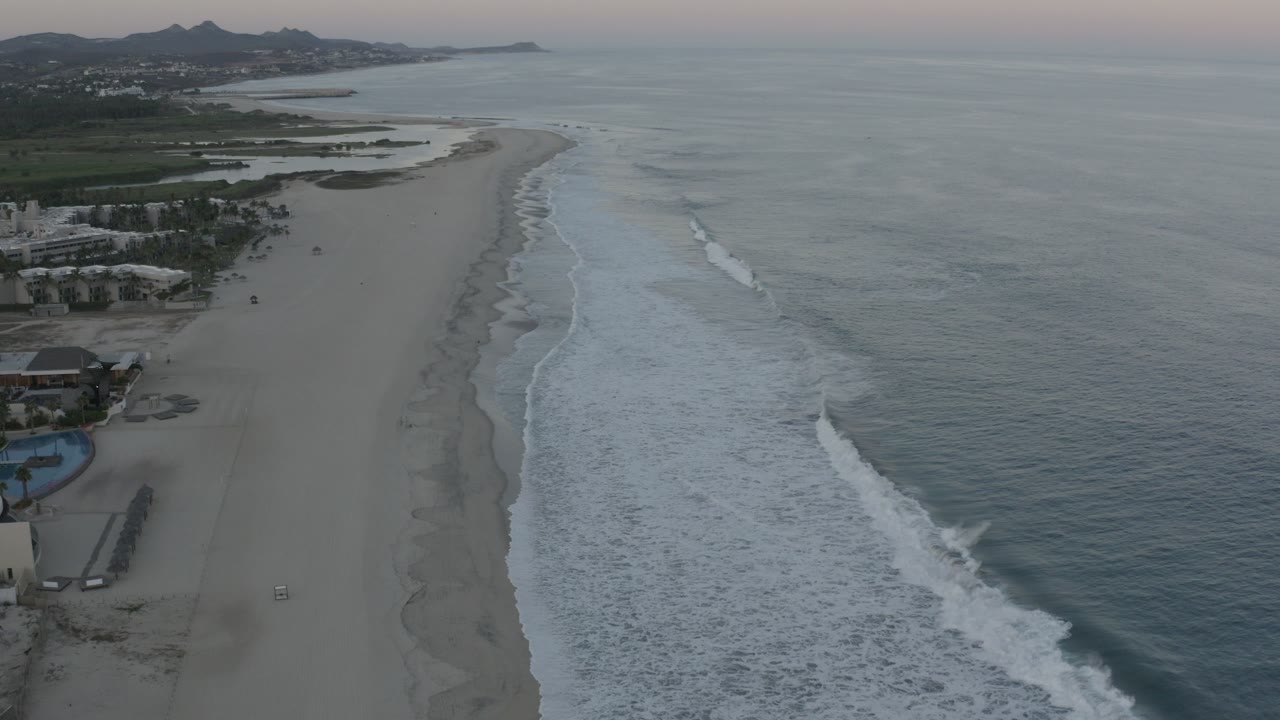 High aerial view over a Los Cabos beach at the start of the clip, with white surf on the right, broad sand down the center, coastal buildings on the left, distant hills on the horizon, and tiny visible people.