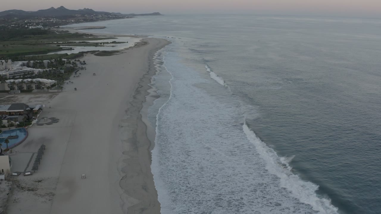 Aerial view over the Los Cabos shoreline one second in, with the beach curving toward the distance, surf bands shifting along the right side, buildings at the lower left, and tiny visible people on the sand.