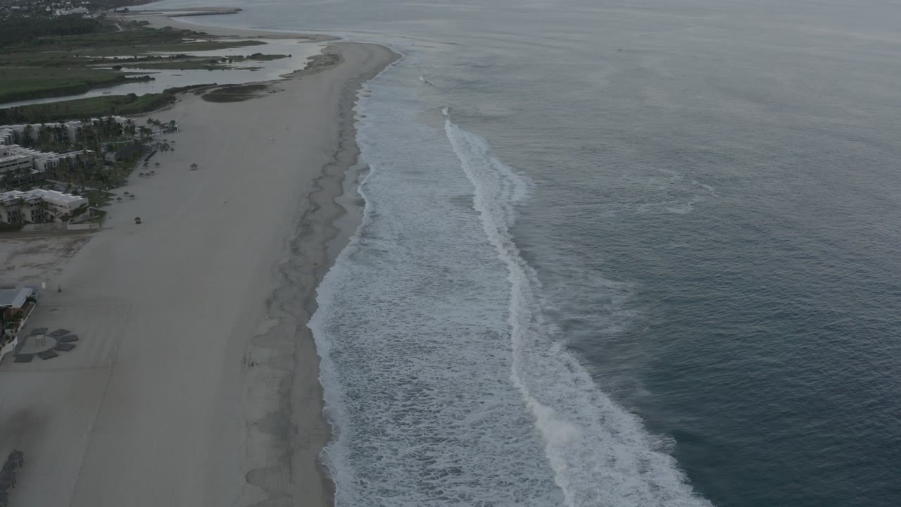Oblique aerial view over a Los Cabos beach with gray-blue ocean on the right, white surf running along the sand, beachfront buildings at left, and no visible people.