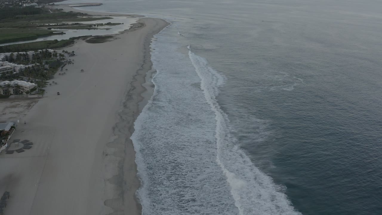 High coastal view in Los Cabos with the beach stretching upward through frame, a bright surf band near center, open water to the right, and no visible people.