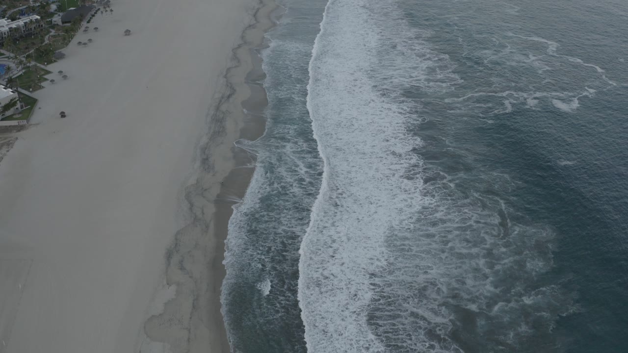 High aerial side view of a Los Cabos beach with pale sand on the left, white foam waves through the center, blue-green ocean on the right, and no visible people.