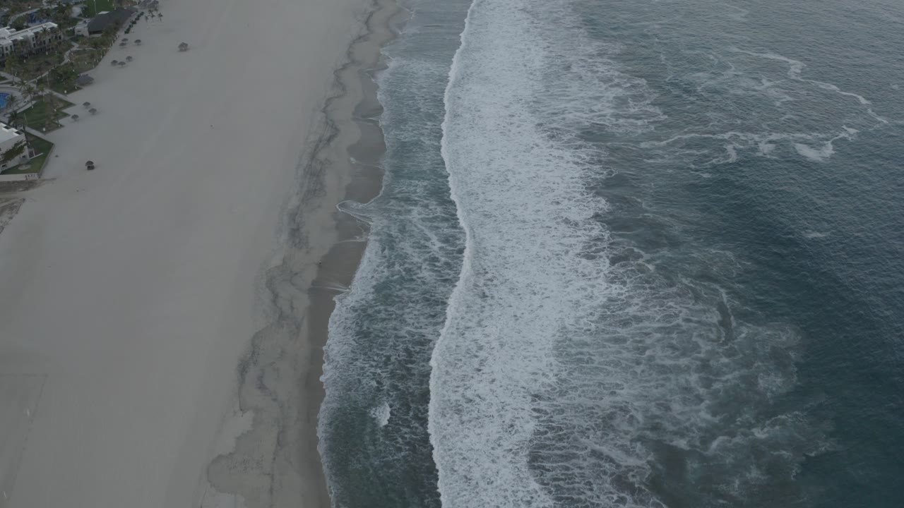 Aerial frame over Los Cabos shoreline showing a wider band of white foam against dark water, smooth pale beach, small beachfront structures near the top edge, and no visible people.