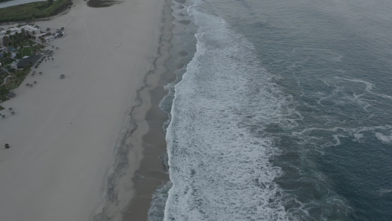 Overhead Los Cabos shoreline frame with foamy waves crossing diagonally, a wide stretch of sand on the left, ocean texture on the right, and no visible people.