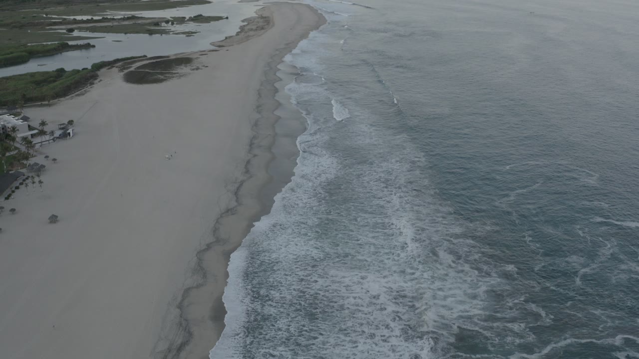 A high aerial view over a Los Cabos beach shows pale sand curving beside blue-gray water, white surf, green vegetation, and no visible people.