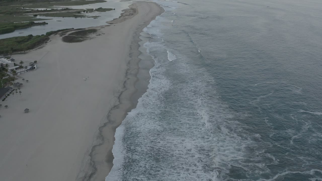 The next aerial frame keeps the Los Cabos shoreline centered, with foamy waves advancing along the beach and dark tidal pools near the vegetation, no visible people.