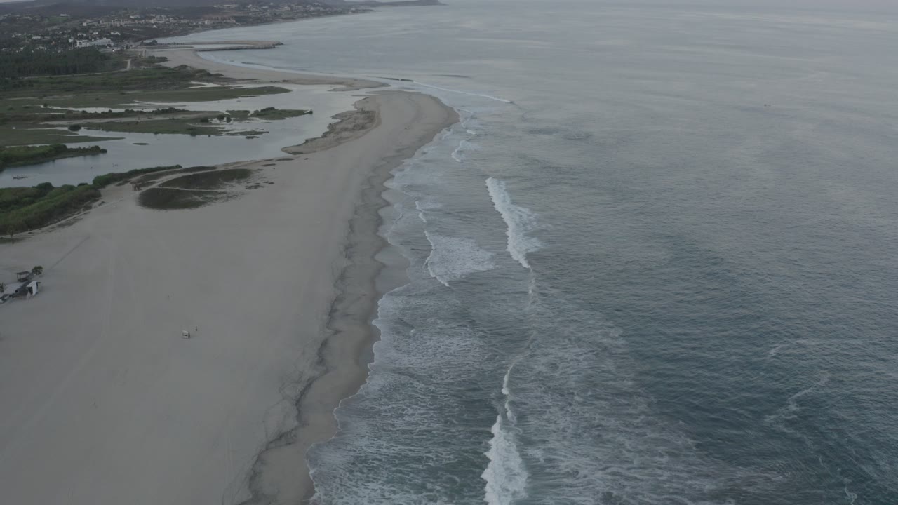 Aerial view of a long beach with waves breaking beside gray-blue ocean.