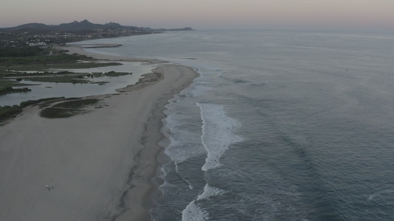 Aerial view of sandy beach, ocean surf, distant hills, and pastel sky.