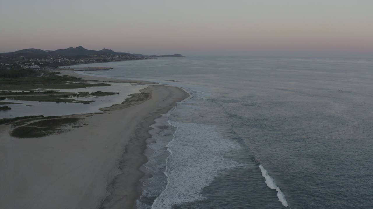 The Los Cabos shoreline appears from above with white surf lines running beside a curving sandbar, darker inland water on the left, and no visible people.