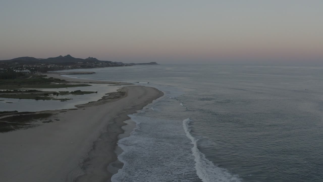 Aerial view over a curved Los Cabos beach at dusk, with white waves on the right, darker lagoon shapes on the left, distant hills, and no visible people.