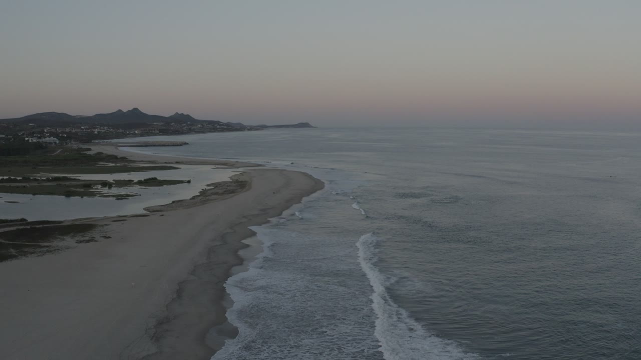 Wide aerial coastline with surf lines and distant hills under evening sky.