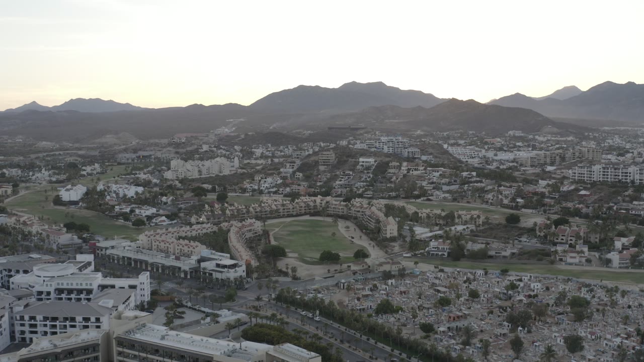 Aerial Los Cabos frame looking across a cemetery foreground toward a green field, dense low-rise buildings, roads, and dark mountain ridges, with no visible people.