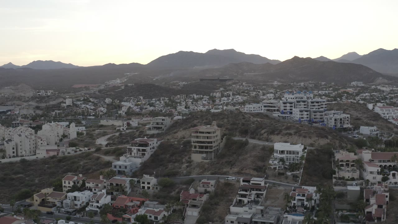 Wide aerial frame over Los Cabos hillside homes and apartment buildings, with roads in the foreground, mountains on the horizon, pale sunrise light, and no visible people.