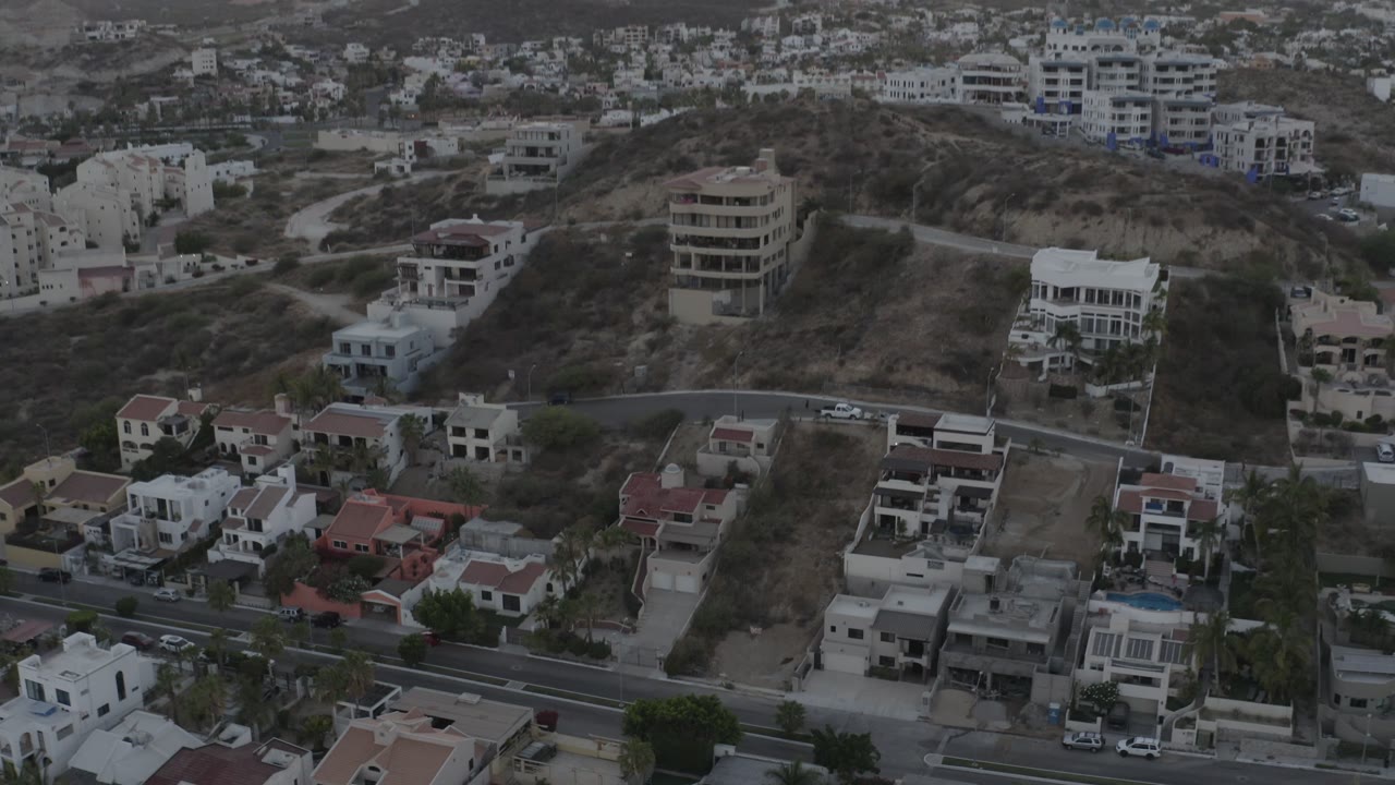 A Los Cabos overhead view shows a paved road at the bottom, compact private homes, graded hillside parcels, sparse vegetation, and no visible people.