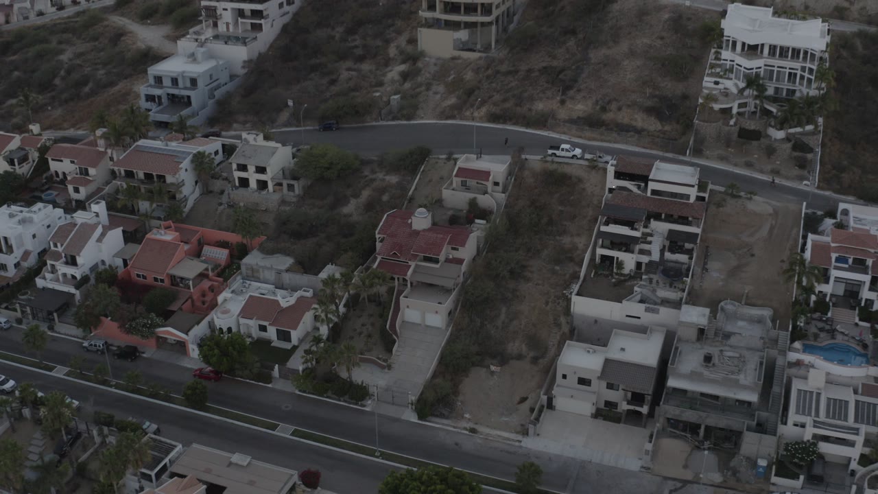 A drone view over Los Cabos, Mexico shows white and red-roofed homes along a curved road, dry hillside lots, parked vehicles, and no visible people.