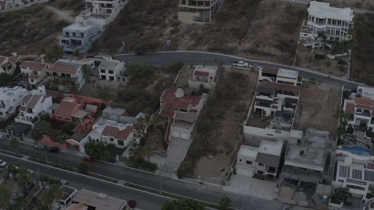 A high aerial frame in Los Cabos, Mexico shows a residential street crossing below hillside homes, with red roofs, white walls, dry lots, and no visible people.