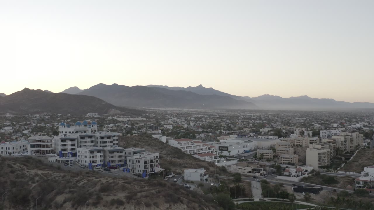 Wide aerial view of hillside buildings, city blocks, and mountains at evening.