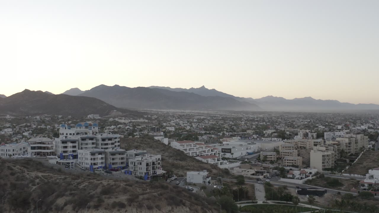 Los Cabos, Mexico aerial frame with a white multi-story building on the left hillside, low city development in the center, layered mountains in the background, and no visible people.