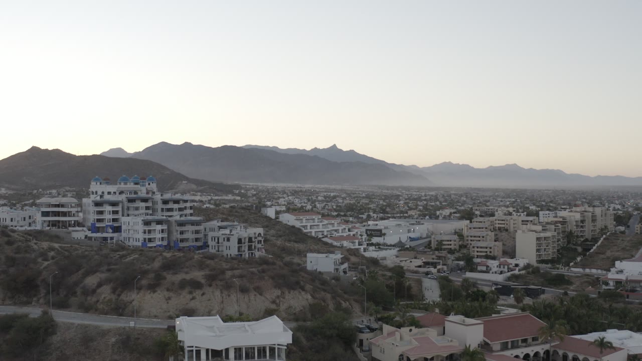 Opening frame of a Los Cabos hillside neighborhood with white homes in front, a broad residential valley beyond, distant mountains, muted light, and no visible people.