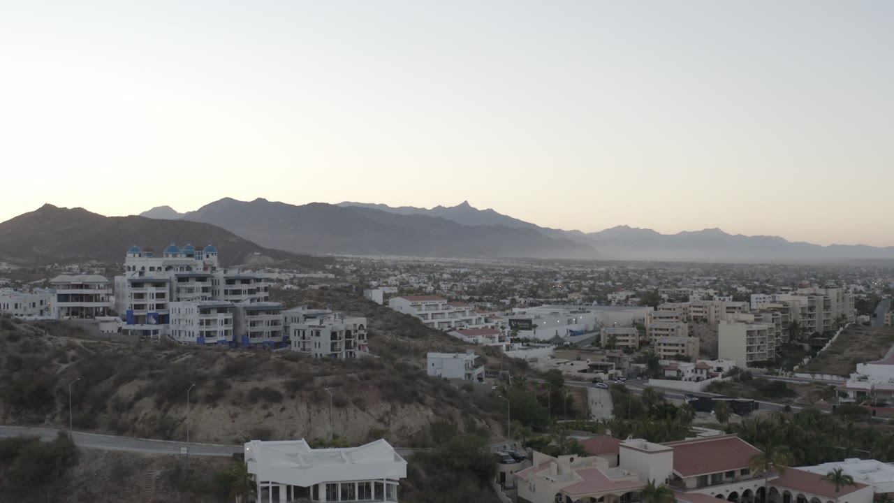 At one second, the Los Cabos view holds on white hillside buildings, low residential blocks across the valley, layered mountains, soft sky, and no visible people.