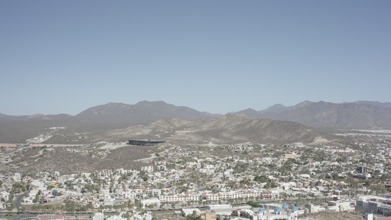Opening frame of San Jose del Cabo with white low-rise buildings below dry hills, hazy mountains along the horizon, and no visible people.