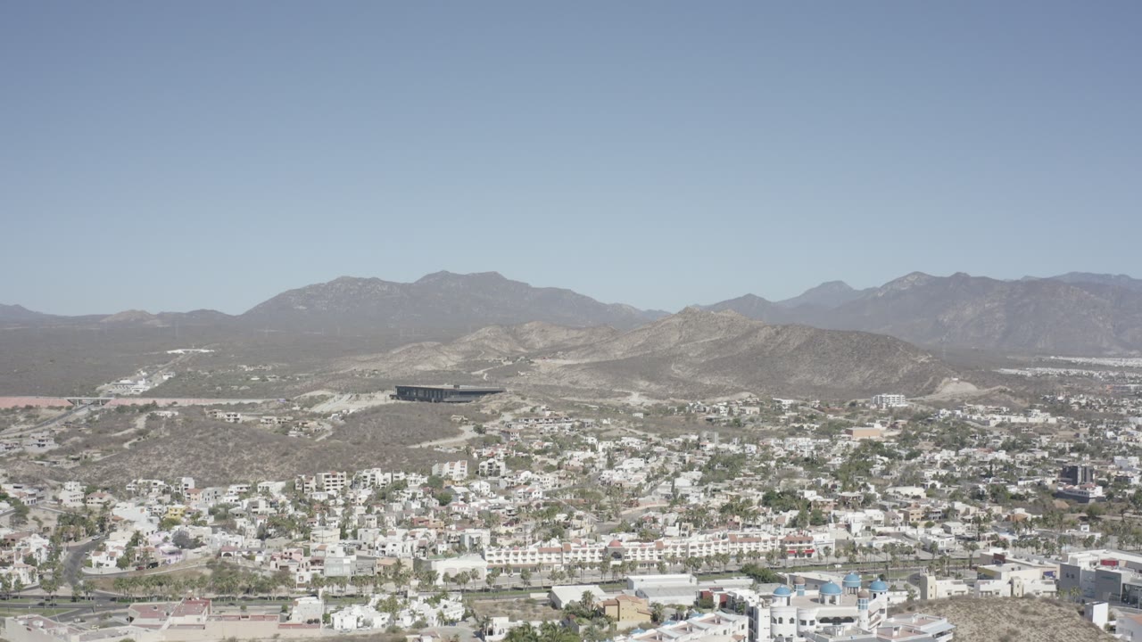 Wide aerial view of white buildings, dry hills, and distant mountains under a pale sky.