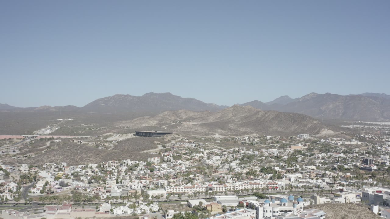 Second aerial frame over San Jose del Cabo, Mexico, with more of the hillside neighborhood visible beneath the wide mountain backdrop and no visible people.