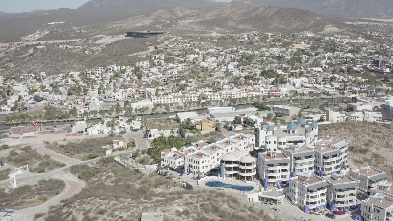 Aerial view of hillside homes, white buildings, pool, roads, and mountains.