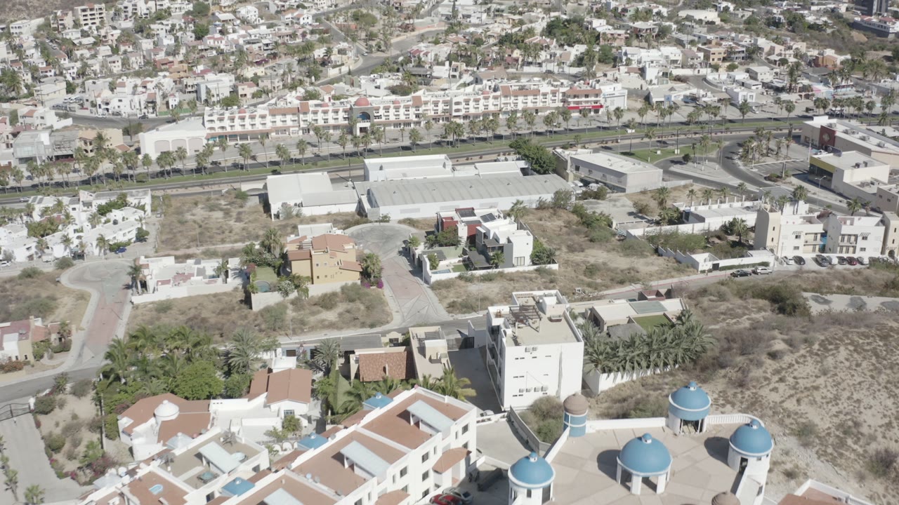 Early frame of San Jose del Cabo showing a high view over white houses with blue roof domes in the foreground, dry lots, roads, apartment blocks, and no visible people.