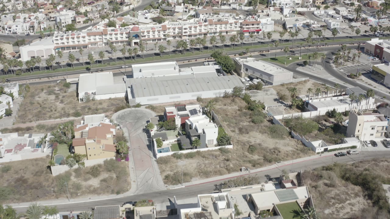 Top-down aerial view over San Jose del Cabo showing white flat-roofed buildings, dry open lots, a curving side street, and no visible people.