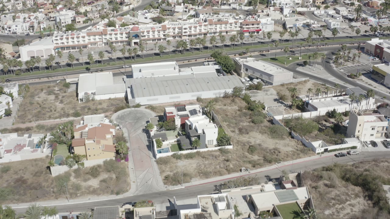 Aerial frame in San Jose del Cabo with a divided boulevard across the upper frame, white low-rise buildings below it, dry lots, and no visible people.