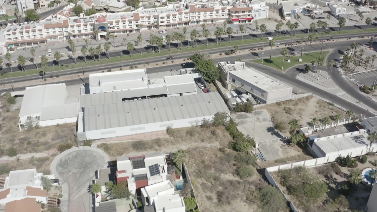 High-angle San Jose Del Cabo frame with box-type white buildings in the foreground, a curved road junction to the right, dry ground, parked vehicles, and no visible people.