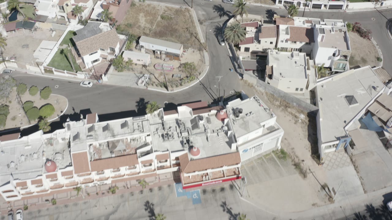 High aerial view over San Jose Del Cabo homes with curved asphalt streets, flat roofs, palms, dry lots, parked cars, and no visible people.