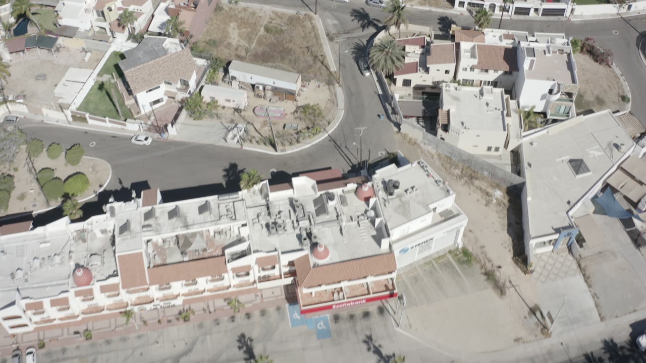 Birdseye frame of San Jose Del Cabo residential blocks, showing villas, a small rounded street junction, palms, dry ground, parked cars, and no visible people.