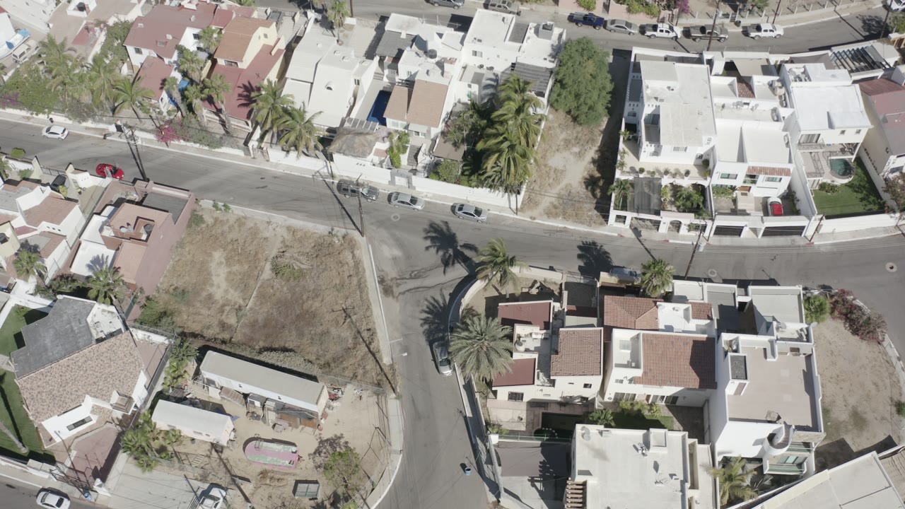 Overhead drone-style view of a San Jose Del Cabo residential block with white modern homes, curved streets, palm trees, parked cars, dry lots, and tiny visible people on the road.