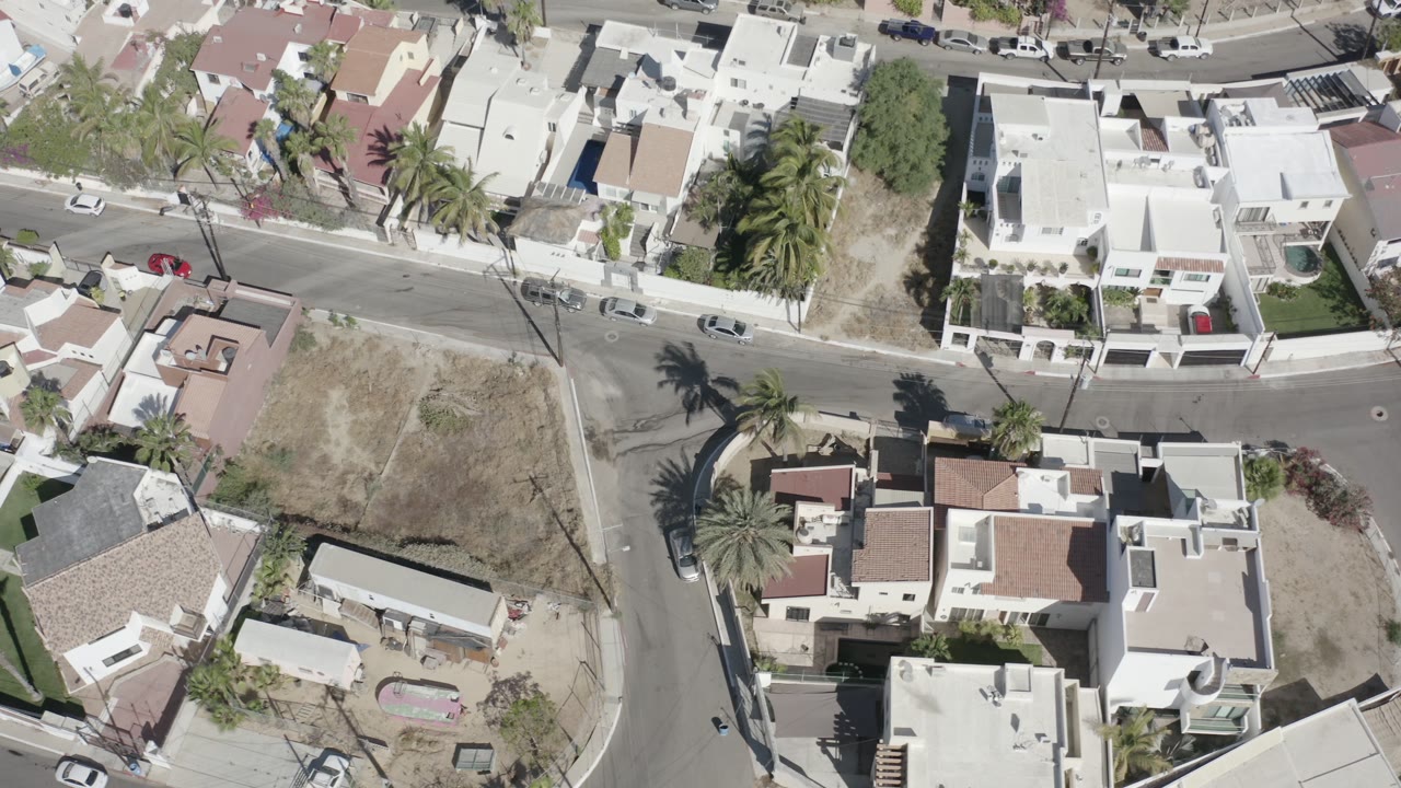 High overhead view in San Jose Del Cabo with a white walled house near the center, palm shadows across the road, parked cars, dry lots, and tiny visible people on the street.