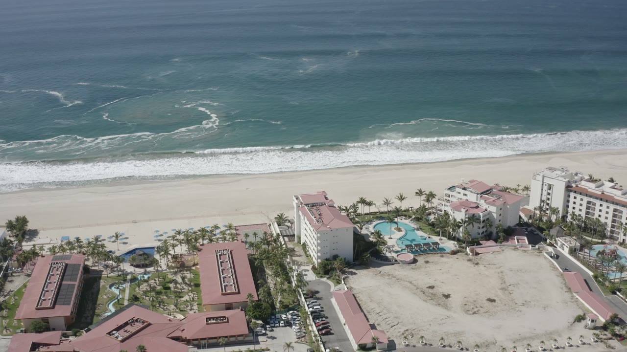 Aerial frame over San Jose Del Cabo, Mexico showing teal ocean, white surf, pale beach, palm-lined resort buildings, turquoise pools, and no clearly visible people.