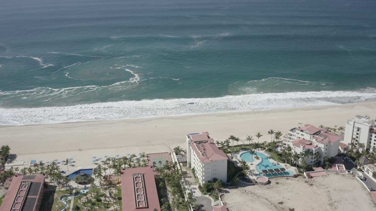 High aerial view of San Jose del Cabo ocean surf meeting a wide sandy beach, with palm-lined resort buildings and pools at the lower edge and no visible people.