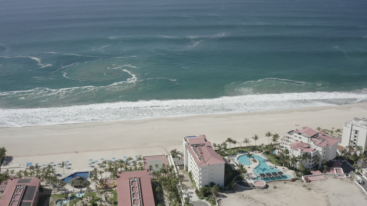 Aerial coastline view with surf lines, pale sand, palms, and resort structures.