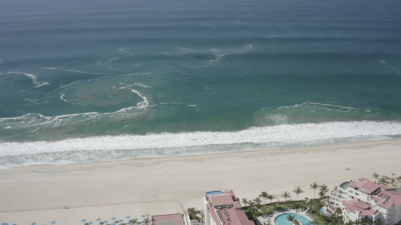 San Jose Del Cabo coastline from above, showing white surf pushed higher onto the sand while pink-roofed beachfront buildings and palms sit near the lower frame edge.