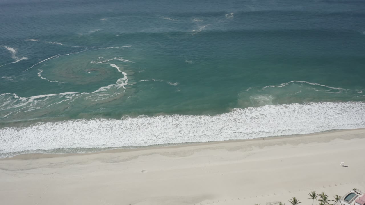 Aerial frame over San Jose Del Cabo coastline with a broad white wave band crossing the center and empty sand below, with no visible people.