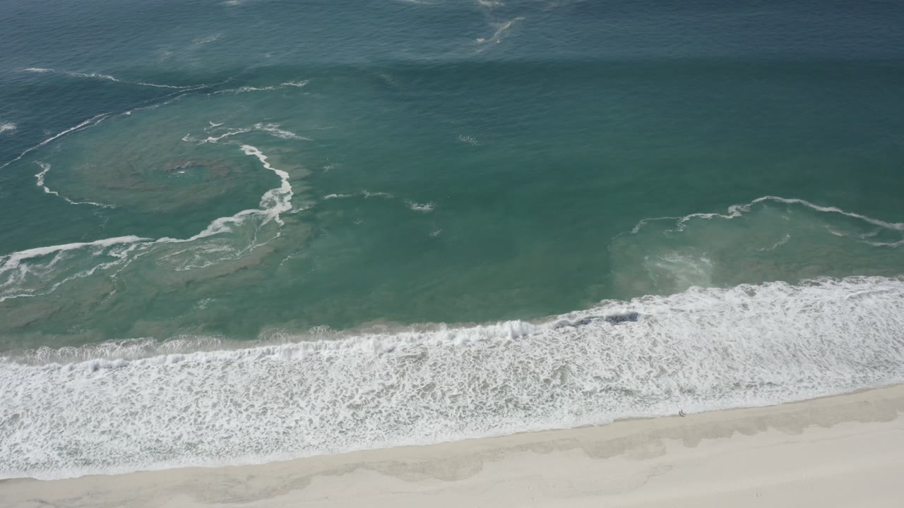 Overhead view of turquoise-green water and white surf rolling toward pale sand in San Jose del Cabo, Mexico, with no visible people.