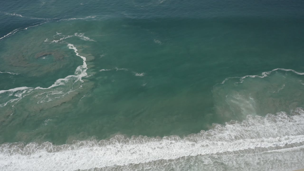 Overhead view of green-blue surf water in San Jose Del Cabo, with a white foam band along the lower frame and no visible people.