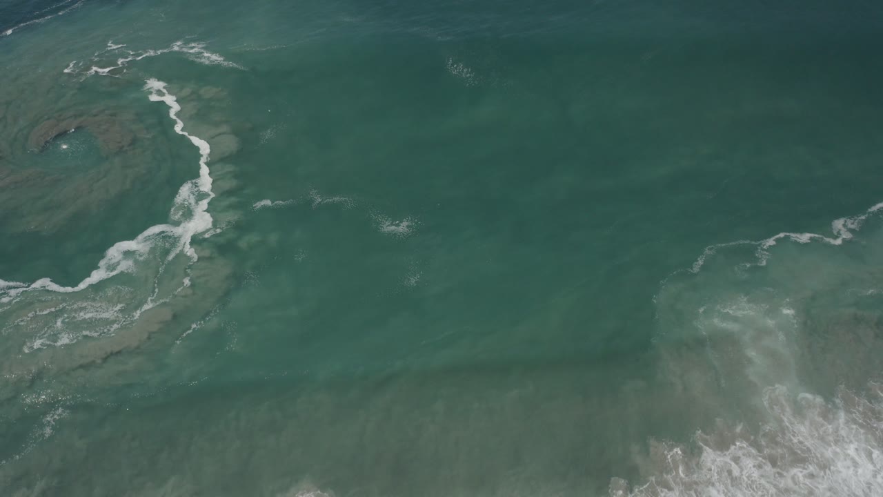 Overhead view of green ocean water in San Jose Del Cabo with white foam tracing a curved line over darker submerged reef shapes, no visible people.