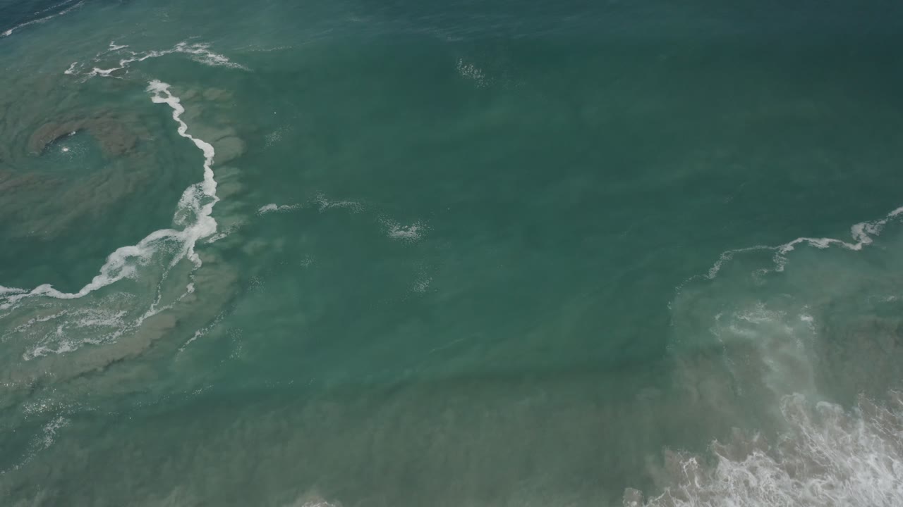 A white surf streak bends through green water off San Jose Del Cabo, with darker underwater rock shapes near the upper left and no visible people.